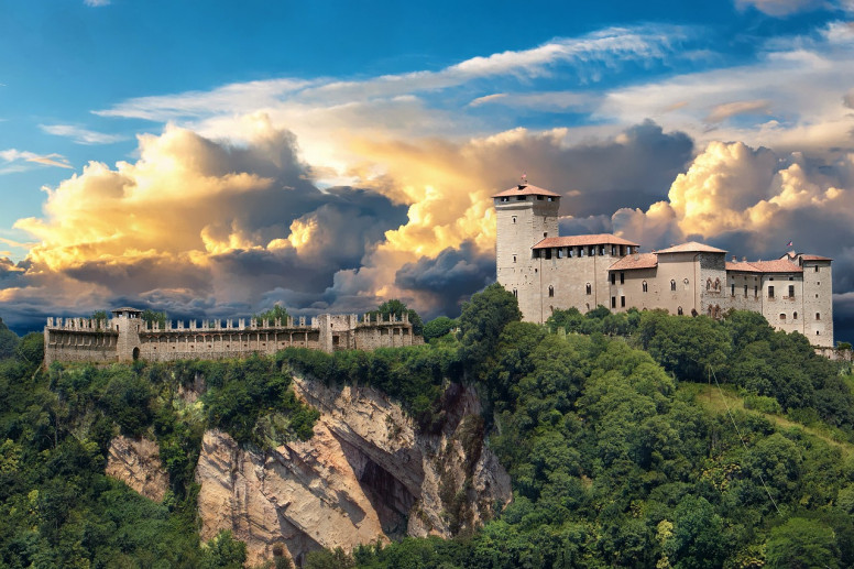 Vista della Rocca di Angera posta sulla sommità di uno sperone di roccia. Il tramonto colora le nuvole di arancione in un cielo azzurro. La Rocca è circondata da piante verdi.