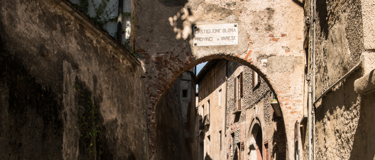 View of a stone arch marking the entrance to the historic center of Castiglione Olona, with a narrow paved street and ancient buildings made of stone and brick.