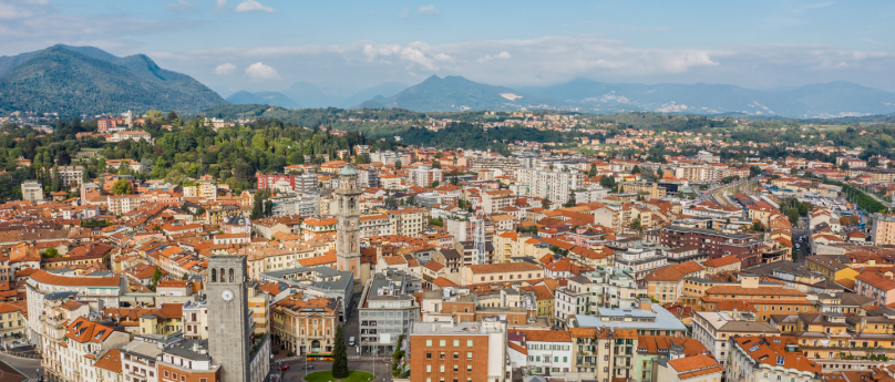 Una vista aerea della città di Varese, con i suoi tetti rossi e grigi che si mescolano tra i palazzi storici e moderni. Al centro si intravede la piazza principale, circondata da alberi verdi. In lontananza, le montagne completano il panorama, creando un contrasto suggestivo con l'urbanità della scena