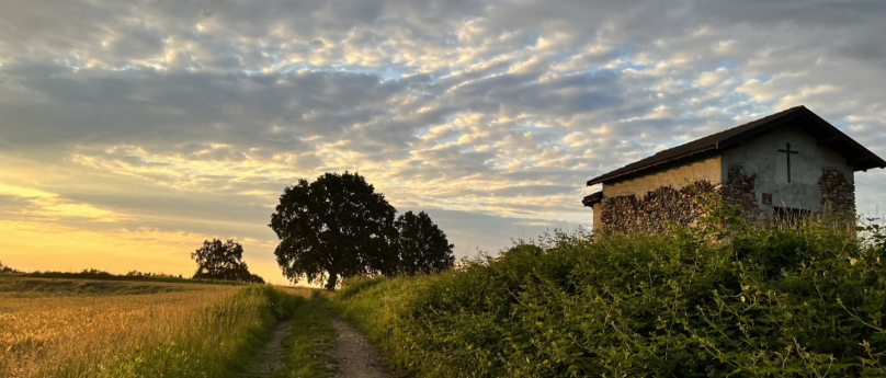 A dirt path crosses a rural landscape at sunset, surrounded by golden fields and green vegetation. To the right, there is a small stone chapel with a cross on its facade, while to the left, large trees can be seen under a cloudy sky illuminated by the warm colors of the sunset. The atmosphere is peaceful and serene, with the contrast between the golden light and the lengthening evening shadows.