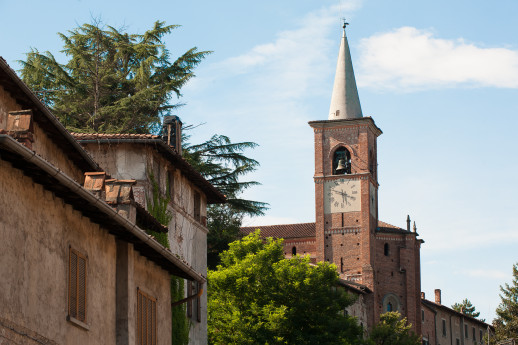 Vista del campanile in mattoni della chiesa di Castiglione Olona con l'orologio, circondato da antiche case e alberi verdi, in un cielo sereno.