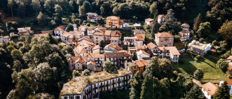 Immagine panoramica di un piccolo villaggio dipinto situato in mezzo alla natura, circondato da una fitta vegetazione. Gli edifici hanno tetti di tegole rosse e sono disposti in modo compatto lungo dolci pendii, creando un'atmosfera pittoresca e tranquilla.