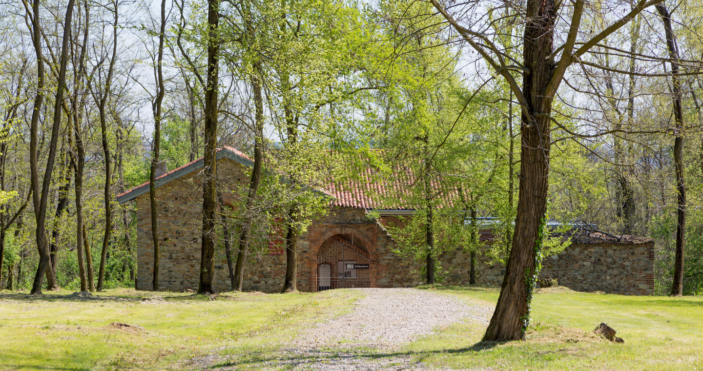 L'immagine mostra un edificio in pietra immerso in un ambiente boscoso, circondato da alberi alti e verdi. Il tetto è coperto da tegole rosse, mentre l'entrata centrale, un arco in mattoni, è chiusa da un cancello di ferro. Davanti all'edificio si estende un sentiero di ghiaia che conduce verso la struttura, creando un'atmosfera tranquilla e isolata, in armonia con la natura circostante.