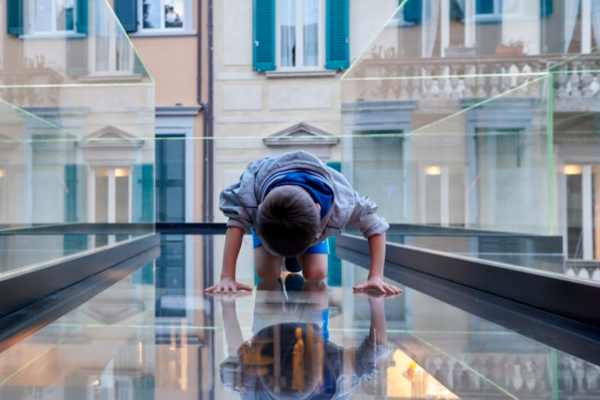 Bambino esplora una passerella di vetro sospesa durante la Varese Design Week, riflettendosi sul pavimento trasparente con edifici residenziali sullo sfondo.