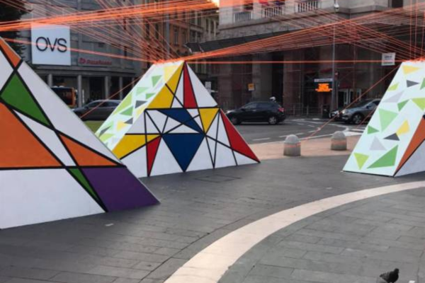 Installazioni geometriche e piramidi colorate esposte in una piazza di Varese per la Varese Design Week, in un contesto urbano con edifici e auto sullo sfondo.