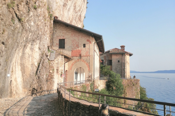 Strutture in pietra costruite nella roccia che creano un passaggio con archi. Il percorso è delimitato da un muretto ed è a picco sul lago Maggiore.